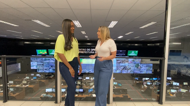 Fotografia. Ambiente interno. As professoras do Rioeduca na TV, Aline Menezes e Cássia Lecce, conversam em frente ao painel de controle do COR - Centro de Operações Rio.