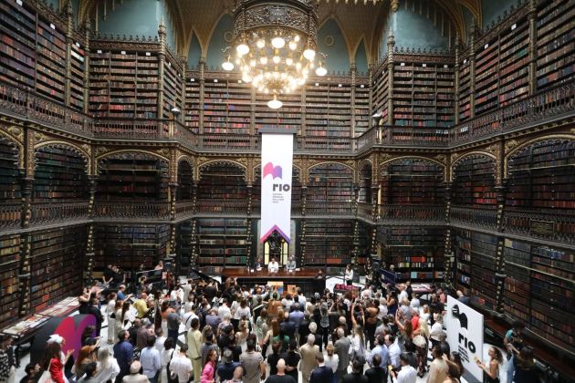 Foto. Dezenas de pessoas no piso térreo de uma grande biblioteca. Pendurado no segundo um banner com a marca do Rio Capital Mundial do Livro 2025.