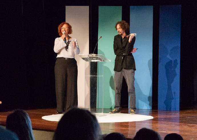 Uma mulher e um homem, ambos com microfone na mão, em um palco de teatro. Em primeiro plano, silhueta de pessoas sentadas na plateia.