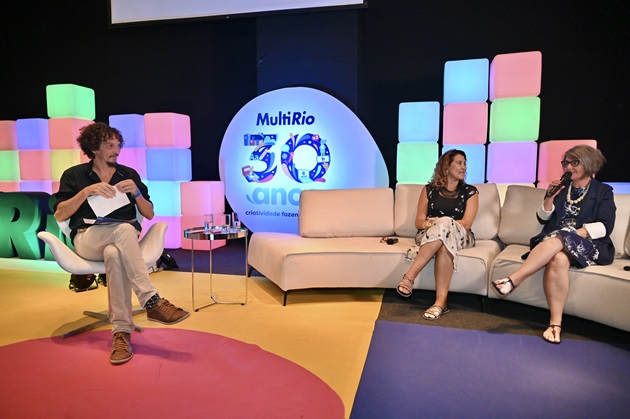 À esquerda, homem sentado em uma cadeira. À direita, sofá com duas mulheres. Atrás, cenário com cubos coloridos e o logotipo MultiRio 30 anos.