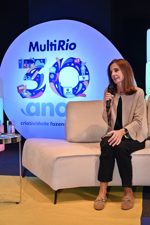 Mulher branca sentada em um sofá com um microfone na mão. Atrás, logotipo MultiRio 30 anos.