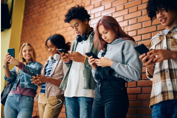 Fotografia. Cinco adolescentes, dois meninos e três meninas de diferentes tons de pele, estão em pé, lado a lado, em frente a uma parede de tijolos. Todos estão com celulares nas mãos. Uma menina posiciona o celular como se estivesse tirando uma selfie, os demais adolescentes estão olhando para as telas.