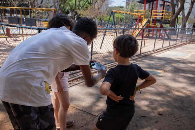 Fotografia. Um adolescente mostra conteúdo em um celular para duas crianças. Eles estão em um parque.