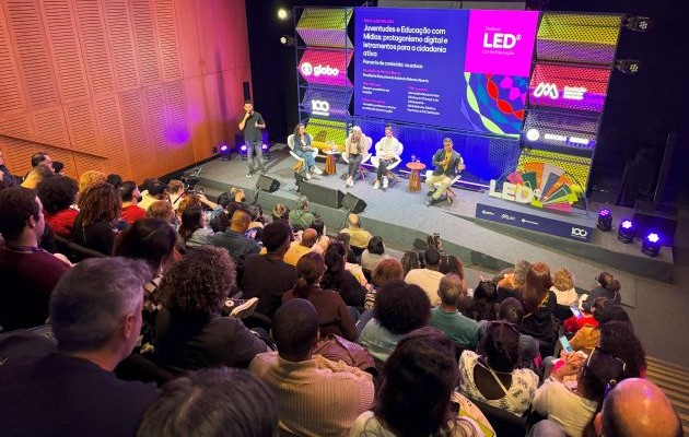 Foto. Em primeiro plano plateia de costas. Ao fundo, palco com palestrantes.